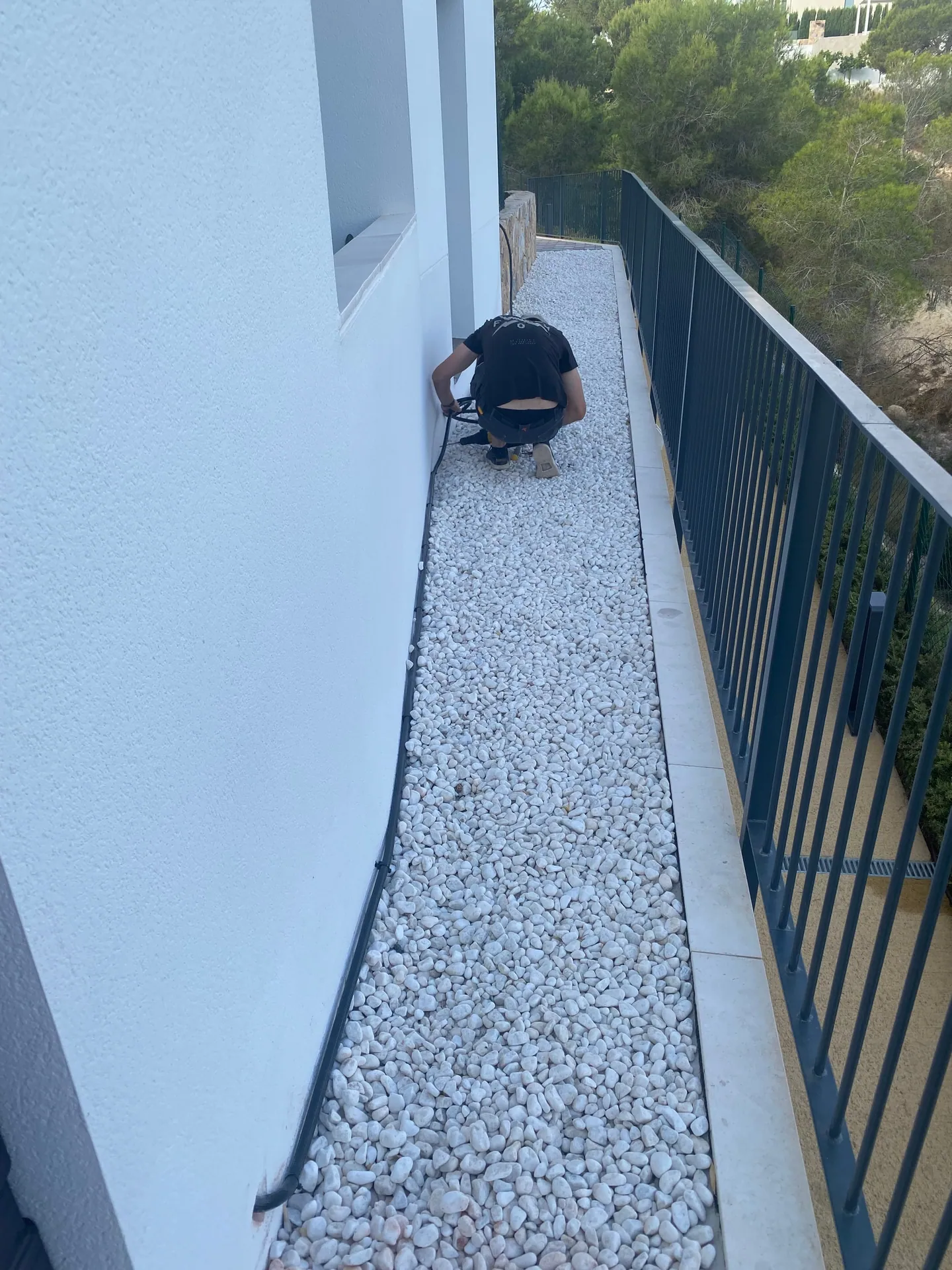 Electricista agachado pasando cable por una franja de grava blanca entre la fachada y la barandilla