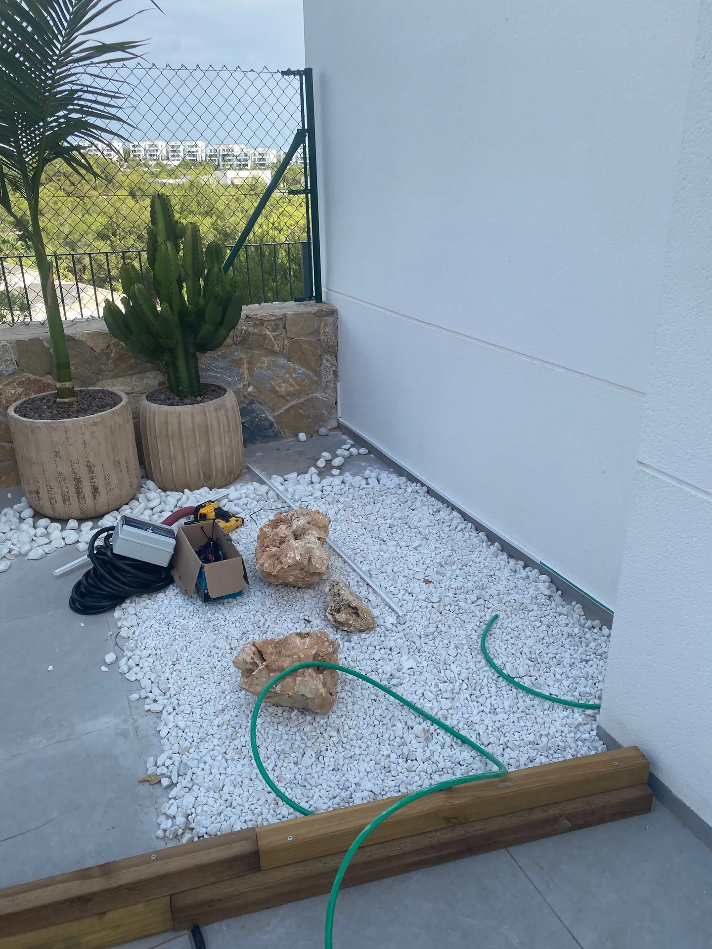Rincón de grava blanca con un rollo de cable y una caja eléctrica preparados junto a las macetas